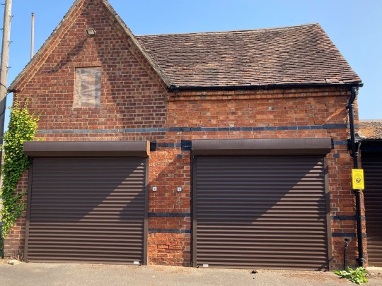 Frontage of garages connected to existing building featuring 2 brown garage doors and 1 boarded up window above door 1.